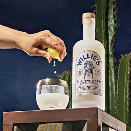 A hand squeezing lime into a glass of Willie's Remedy+ 5 mg THC Social Tonic next to the 750 ml bottle