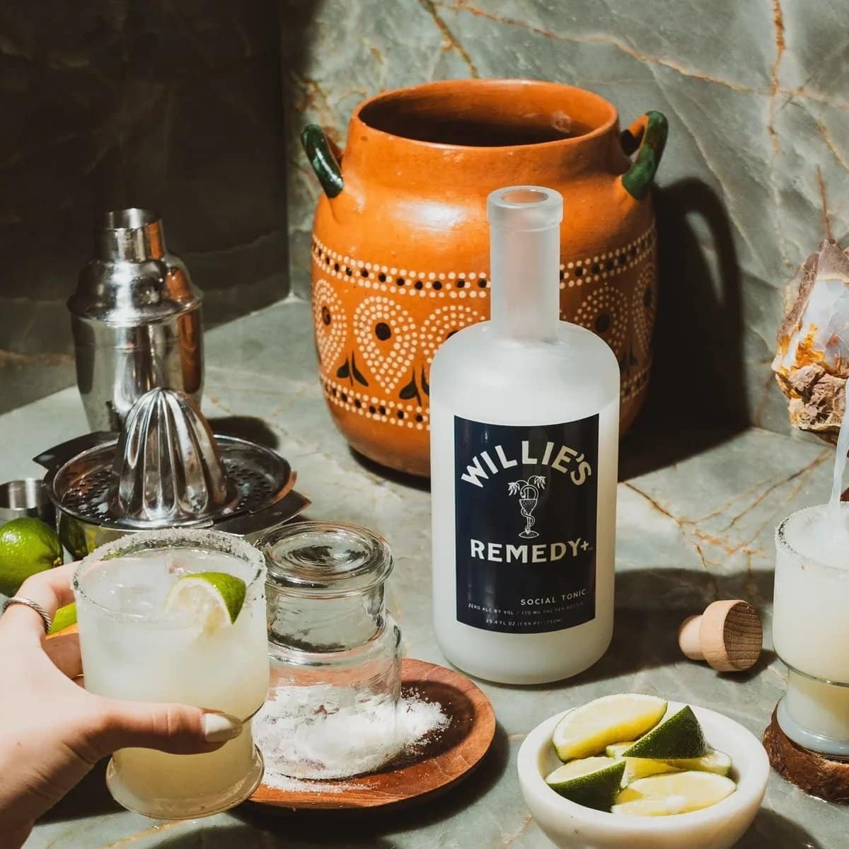 Willie's Remedy+ 750 ml Social Tonic bottle on a bar with a shaker, juicer, limes and a salt-rimmed glass